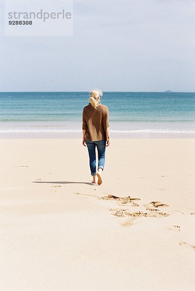 Eine Frau  die barfuß am Strand spazieren geht und dabei Fußspuren im Sand hinterlässt.