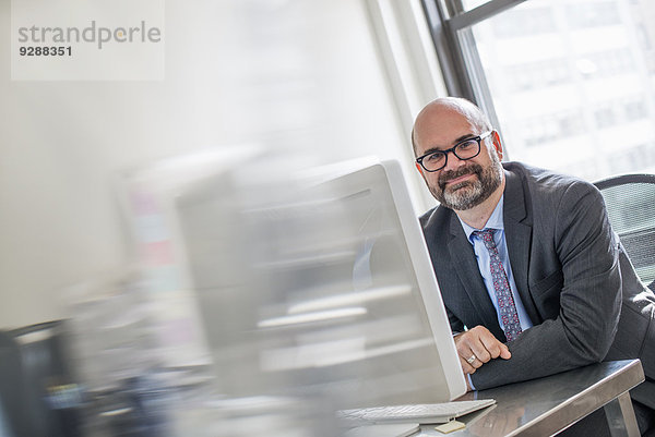 Büroalltag. Ein Mann in Anzug und Krawatte sitzt an seinem Schreibtisch und sieht sich am Computer um.