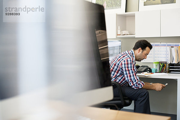 Ein Mann  der in einem Büro sitzt und sein Smartphone kontrolliert.