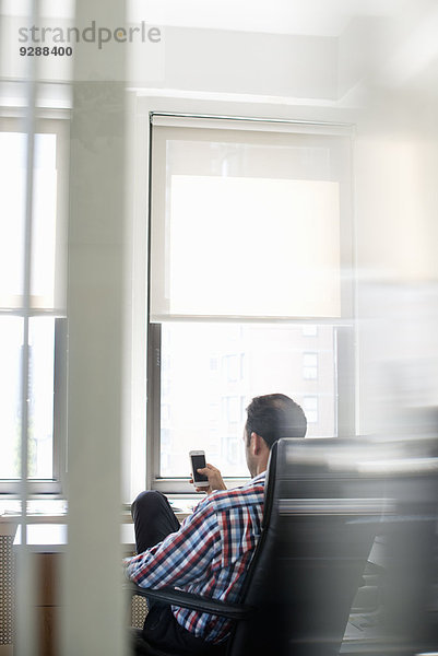 Ein Mann  der in einem großen Bürostuhl sitzt  sich zurücklehnt und sein Smartphone kontrolliert.