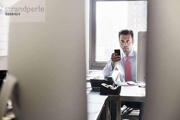Ein Mann überprüft sein Smartphone und setzt sich an seinen Büroschreibtisch.