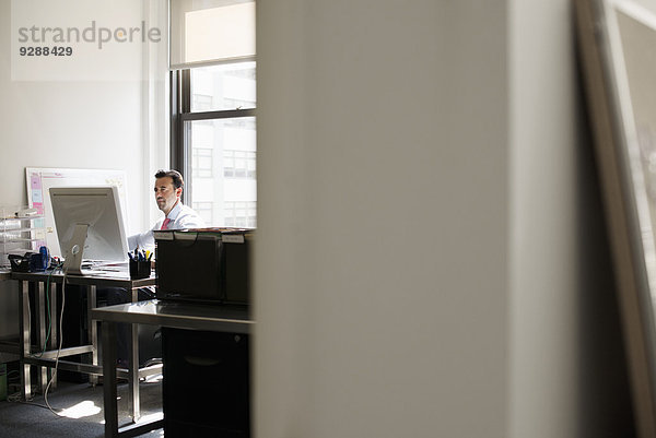 Ein Mann  der an einem Schreibtisch in einem Büro sitzt und einen Computer benutzt.