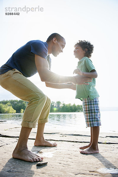 Ein Mann hebt einen kleinen Jungen hoch  der an einem See in der Sonne spielt.