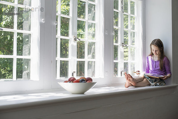 Mädchen  das am Fenster sitzt und ein Buch liest.