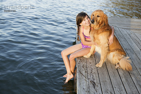 Ein Mädchen und ihr Golden-Retriever-Hund sitzen auf einem Steg an einem See.