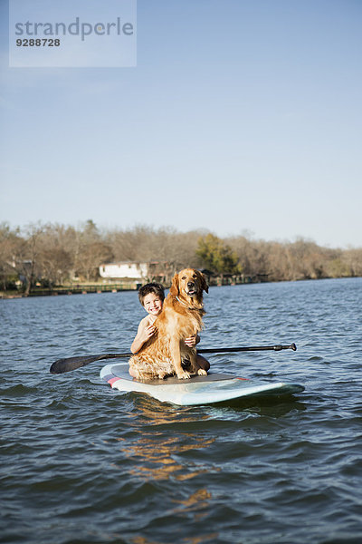 Ein Kind und ein Apportierhund sitzen auf einem Paddelbrett auf dem Wasser.