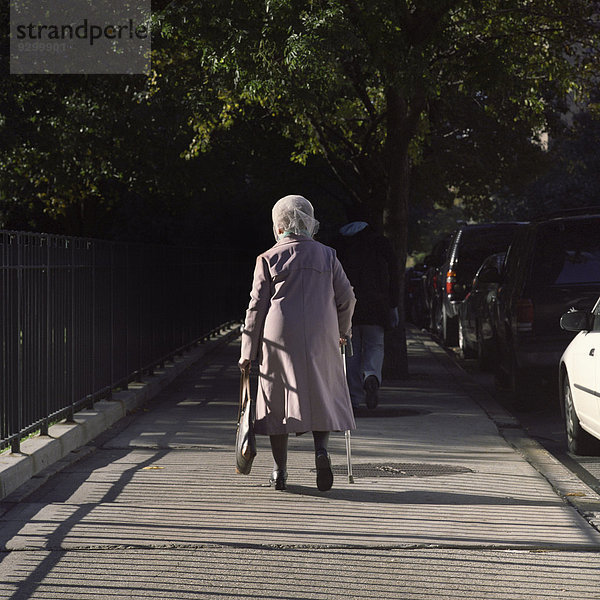 Eine ältere Frau mit einem Stock auf einem Bürgersteig  Rückansicht