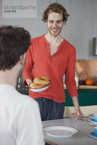 Lächelnder schwuler Mann mit Croissants auf dem Teller  der den Partner zu Hause ansieht.