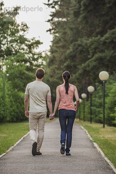 Rückansicht eines jungen Paares  das im Park Hand in Hand geht.