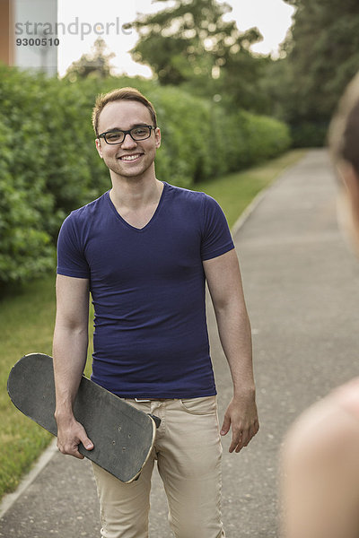 Glücklicher junger Mann hält Skateboard  während er die Frau im Park ansieht.