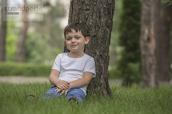 Porträt eines süßen Jungen  der sich gegen einen Baum im Park entspannt.