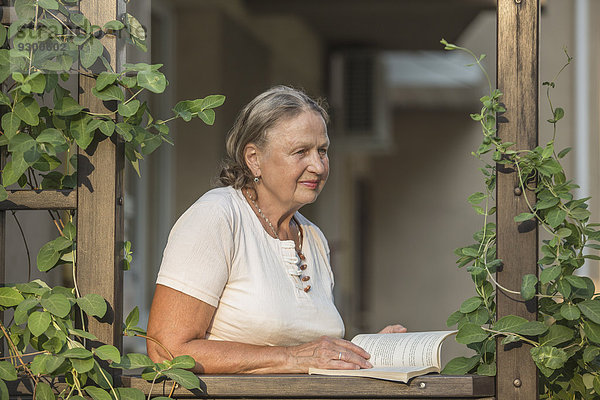 Seniorin hält Buch  während sie auf den Balkon schaut.