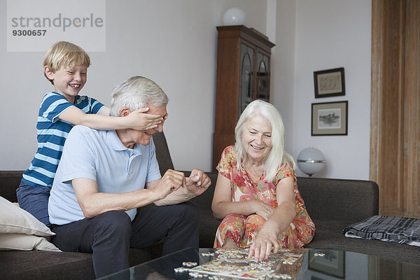 Junge  der die Augen des Großvaters bedeckt  während die ältere Frau ein Puzzle am Tisch im Wohnzimmer arrangiert.