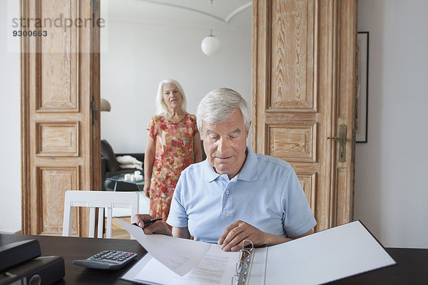 Senior Mann bei der Durchsicht von Finanzdokumenten mit Frau im Hintergrund zu Hause