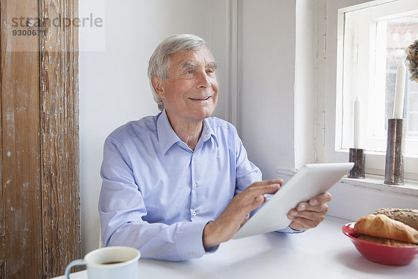 Nachdenklicher älterer Mann schaut weg  während er das digitale Tablett am Tisch hält.