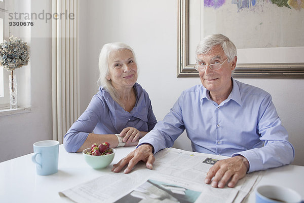 Porträt eines glücklichen älteren Paares mit Zeitung am Tisch