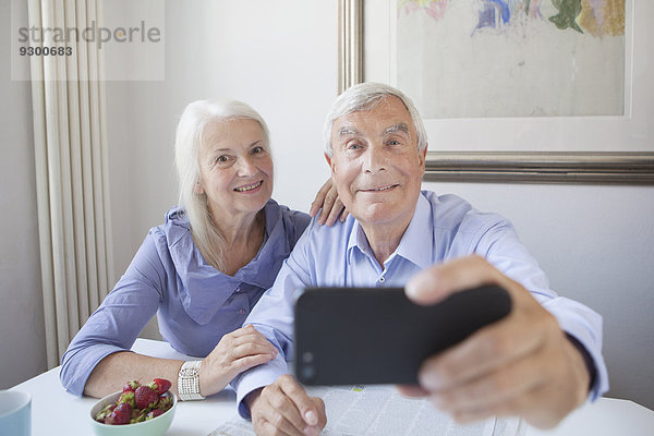 Glückliches Seniorenpaar beim Selbstporträt durch Smartphone am Tisch im Haus