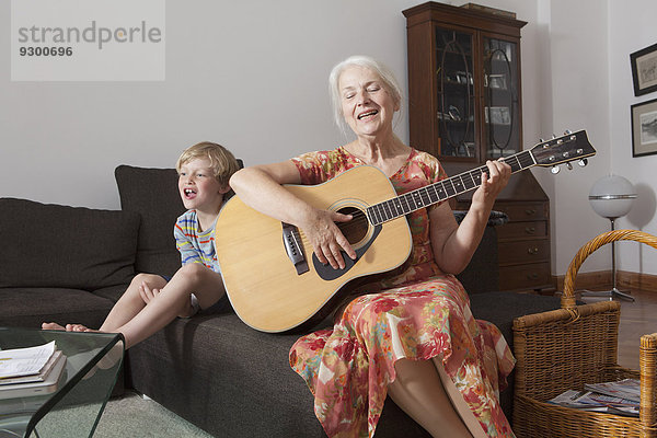 Verspielter Junge sitzend mit Großmutter beim Gitarrespielen auf dem Sofa zu Hause.
