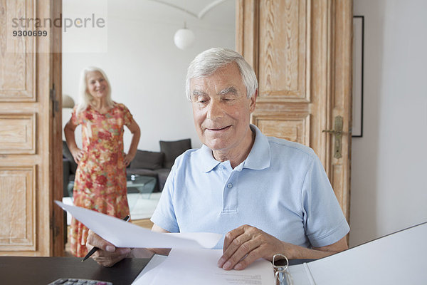 Senior-Mann  der zu Hause mit Frau im Hintergrund Finanzdokumente prüft