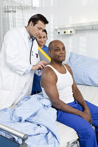Ein Arzt und eine Krankenschwester untersuchen einen Patienten  der auf einem Krankenhausbett sitzt.