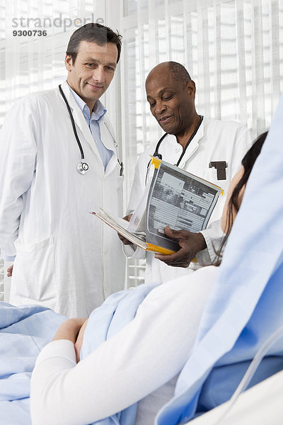 Zwei lächelnde Ärzte beraten einen Patienten  der in einem Krankenhausbett liegt.