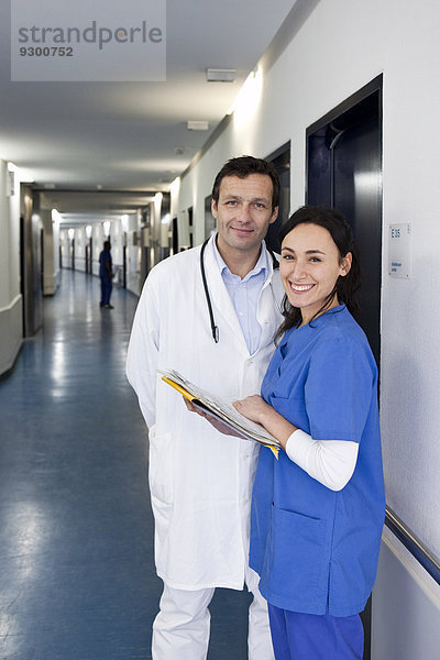 Eine Krankenschwester in Peelings konsultiert einen Arzt in einem Laborkittel über eine medizinische Akte und schaut in die Kamera.