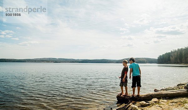 Zwei Jungen stehen auf einem umgestürzten Baum und schauen in den See.