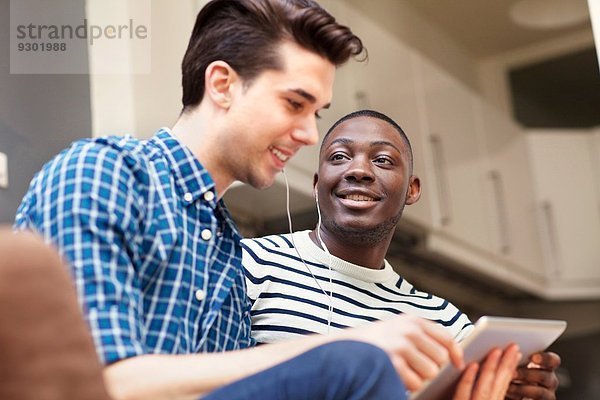Zwei junge Männer hören Musik auf digitalem Tablett auf dem Sofa im Wohnzimmer.