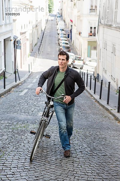 Mittlerer erwachsener Mann schiebt Fahrrad die gepflasterte Stadtstraße hinauf