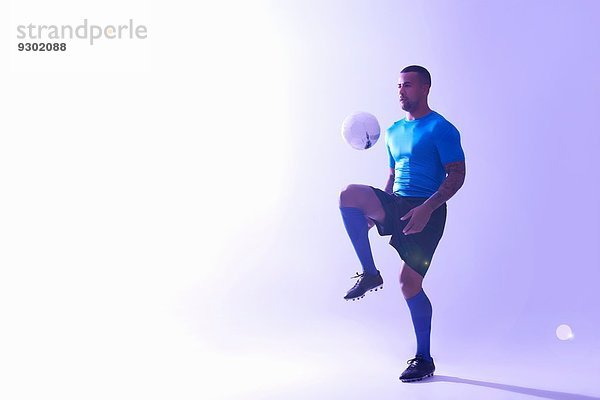 Studioaufnahme eines jungen männlichen Fußballspielers  der den Ball mit dem Knie in der Luft hält.