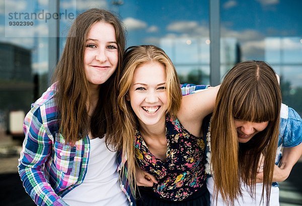 Porträt von drei jungen Frauen  die vor einem gläsernen Bürogebäude lachen.