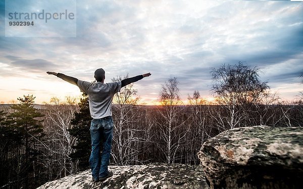 Junger Mann mit ausgestreckten Armen auf einer Felsformation bei Sonnenuntergang