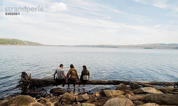Drei Wanderfreunde sitzen in einer Reihe auf einem umgestürzten Baum am Seeufer.