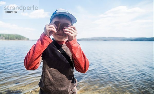 Porträt des schelmischen Jungen mit Keksen über den Augen am Seeufer