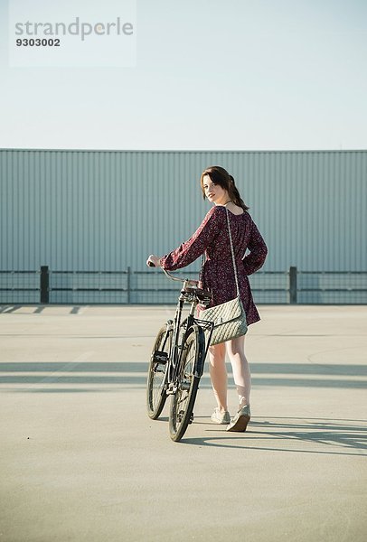 Junge Frau schaut zurück  während sie das Fahrrad auf dem leeren Parkplatz schiebt.