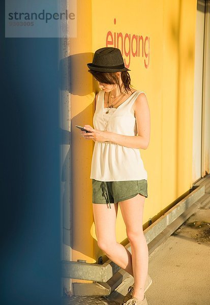 Junge Frau  die sich an die Wand lehnt und auf dem Smartphone SMS schreibt