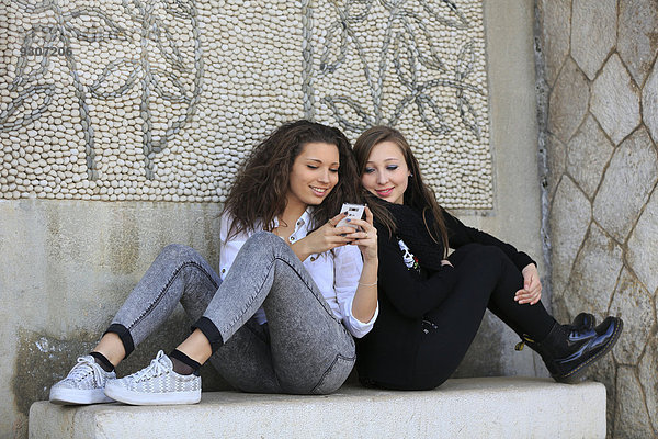 Zwei Freundinnen  Teenager  sitzen Rücken an Rücken auf einer steinernen Bank und lesen eine SMS auf einem Handy  Menton  Provence-Alpes-Côte d?Azur  Frankreich