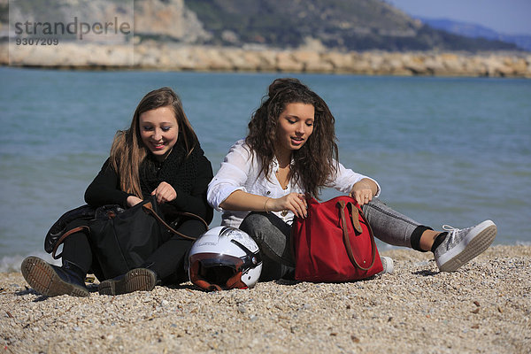 Zwei Freundinnen  Teenager  sitzen am Strand mit Taschen und Sturzhelm  Menton  Alpes-Maritimes  Provence-Alpes-Côte d'Azur  Frankreich