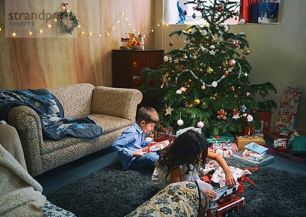 Schwester und Bruder beim Spielen und Auspacken von Weihnachtsgeschenken auf dem Wohnzimmerboden