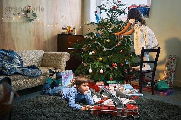 Mädchen arrangiert Weihnachtsbaum  während der Bruder mit Weihnachtsgeschenken auf dem Wohnzimmerboden spielt.