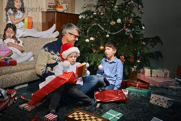 Vater auf dem Wohnzimmerboden öffnet Weihnachtsgeschenke mit Kindern
