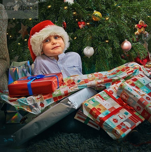 Porträt des süßen Jungen auf dem Boden sitzend mit Weihnachtsgeschenken über ihm