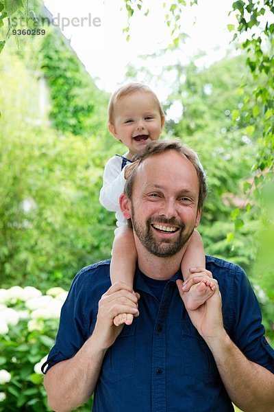 Porträt eines Mädchens  das auf den Schultern des Vaters im Garten sitzt