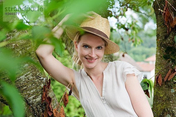 Porträt der mittleren erwachsenen Frau mit Strohhut neben dem Gartenbaum