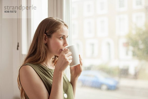 Junge Frau trinkt Kaffee und blickt durchs Wohnzimmerfenster