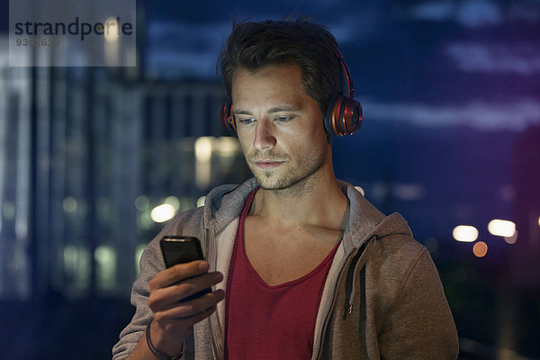 Porträt eines jungen Mannes mit Smartphone und Kopfhörer beim Musikhören bei Nacht