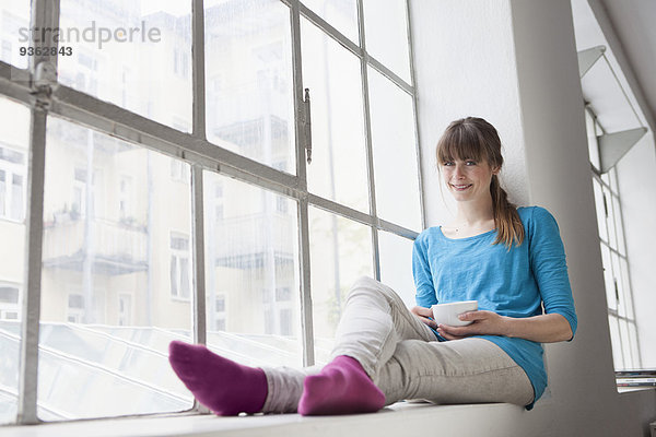Porträt einer lächelnden jungen Frau mit einer Tasse Kaffee auf einer Fensterbank im Büro.