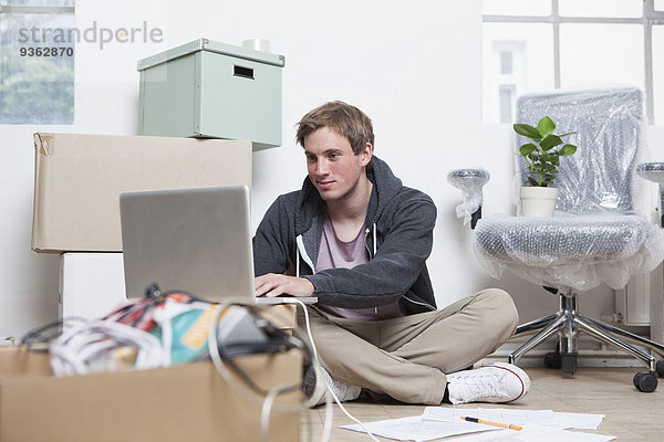 Porträt eines jungen Mannes  der mit seinem Notizbuch zwischen Pappkartons in einem Büro sitzt.