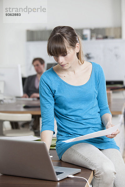 Junge Frau sitzt auf ihrem Schreibtisch in einem Büro mit Laptop
