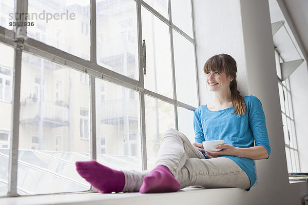 Porträt einer jungen Frau mit Tasse Kaffee auf der Fensterbank im Büro